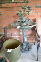 Photo of round metal birdbath with floral fountain, placed against brick wall near a patio chair and a large planter.