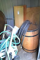 Photo showing wooden barrel with black garden hose coiled over it, plastic bucket, and some boards leaning in background.