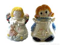 Photo showing both ceramic cookie jars: Angel on left with blonde hair and closed hands, Raggedy Ann on right with red pigtails, apron, and smiling face.
