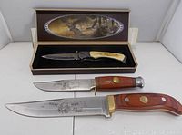 Photo shows three Wild West themed knives: a folding knife in an open box with eagle artwork on lid, two fixed blade knives with wooden handles and incised images and signatures on blades.