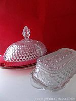 Two pressed glass butter dishes side by side on white surface with red background, one ruby glass base with clear patterned dome lid, one clear rectangular pressed glass butter dish with lid