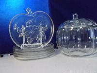 Image showing 5 clear glass apple-shaped snack plates stacked beside a clear glass pumpkin-shaped cookie jar with lid, all placed against a blue background.