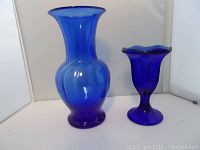 Pair of cobalt blue glass vases shown side by side. Larger vase has a rounded body with flared neck. Smaller vase has scalloped rim and pedestal base.
