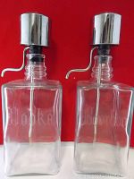 Front view of two clear vintage etched glass liquor decanters labeled 'Vodka' and 'Bourbon' with metal pump dispensers against a red background.