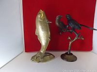 Side view of brass fish sculpture standing next to brass birds sculpture with two birds perched on a branch, both on flat bases, shown against red background.