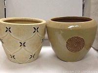 Photo showing two ceramic garden pots side by side on a shelf, one cream with floral cutouts and the other beige with medallion decoration.