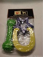 Photo of a sealed 3-piece rubber dog toy combo pack with a green bone-shaped toy, purple star-shaped toy, and yellow horseshoe-shaped toy.