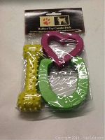 Packaged rubber dog toy combo pack showing yellow textured bone, pink heart-shaped toy, and green horseshoe-shaped toy.