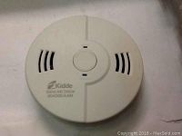 White Kidde smoke and carbon monoxide alarm device photographed from the top, showing its vents, central button, and branding.