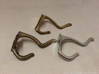 Photo showing three metal coat hanging hooks in brass, bronze, and silver tones with a vintage curved double-hook design.