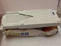 White plastic V Slicer Plus mandoline slicer placed on its original box, showing wear and dirt marks on the slicer and creases on the box.