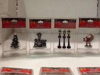 Photo showing four holiday village accessory items still in original plastic packaging: a Christmas tree, a small train, three lamp posts, and a Santa Claus figurine.