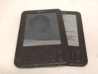 Two black Amazon Kindle e-readers side by side on a white surface, each with physical keyboards and active screens