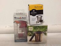 Photo shows three boxed reusable coffee and tea brewing accessories on a shelf against a white wall: Brew & Save reusable Keurig filter, My K-Cup reusable coffee filter in black and yellow box, and Rishi Loose Tea Infuser Basket in green and white box.