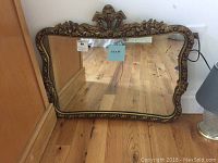 Mirror shown leaning against a wall on a wood floor, showing ornate gold frame and damaged mirror surface.