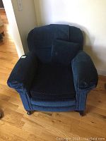 Front view of a blue upholstered armchair with a textured fabric, placed on a wooden floor by a white wall.