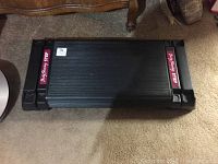 Top-down view of the black rectangular exercise step platform with Body Shaping Step pink labels on either end on carpeted floor near furniture.