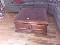 Front view of the square wooden coffee table placed on a hardwood floor in front of a sectional sofa. Shows the tabletop and fronts of two drawers with round knobs.