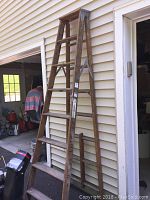 Wooden folding ladder approximately 9 feet tall leaning against garage siding with wide steps and metal hinges at top.