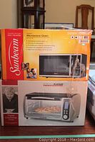 Boxed Sunbeam microwave oven and Gold Ramsey toaster oven stacked, showing model and branding on packaging.