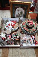 Photo showing two trays with Christmas-themed plates, mugs, glass vase, ceramic lidded container, small decorations, and ornaments.