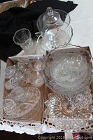 Two trays filled with assorted clear glass and crystal tableware, including bowls, cups, salt and pepper shakers, cream and sugar set, and crystal vase.