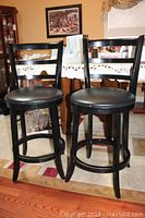 Pair of black wooden bar stools with vinyl cushioned seats shown from front, placed on hardwood and area rug.