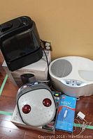 Photo showing the Life digital ultrasonic humidifier, Charlescraft foot massager, Bionaire humidifier, Life heating pad in packaging, and grab bar box on wooden floor near wall outlet.