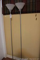 Full view of two tall matching floor lamps with metal shafts and white plastic cone-shaped shades standing side by side against a wall.