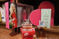 Photo showing boxed Just Me perfume and body lotion set, DKNY Be Delicious perfume bottle and box, heart-shaped pink case, small floral-style perfume bottle, and clear perfume bottle on wooden floor