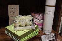 Assorted boxed soaps and fragrances on a wooden shelf, including Issey Miyake boxed soaps, pink rose-shaped soaps in clear box, and Perry Ellis 360° spray