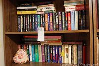 Two wooden shelves filled with rows of assorted paperback books and a heart shaped container on the bottom shelf.