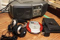 Wide angled photo showing the Panasonic AM FM cassette player, GE portable cassette recorder, Misaki CD player, and three black and silver wired headsets arranged on wooden floor.