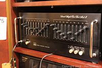 Front view of the ADC Stereo Frequency Equalizer SS 110 Mark II inside a wooden cabinet showing the device front panel with metal handles and sliders.