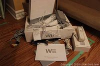 White Nintendo Wii console on stand with connected cables, two Wii remotes (one in box, one loose with jacket), nunchuk, sensor bar, AC adapter and AV cable arranged on floor with manuals.
