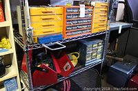 View of metal shelving unit holding two yellow plastic and two metal small parts drawer cabinets, with additional tool boxes and extension cord below.