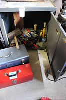 Metal cabinet door open showing hand tools inside along with two metal toolboxes on floor in front.