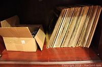 Wooden shelf containing a large number of vintage vinyl records stored vertically next to a cardboard box.