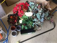 Photo of the assorted faux plants including red poinsettia, ivy, fern, and small cactus. Basket of assorted plastic fruit beside them. Several decorative planters visible on floor.