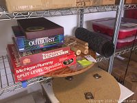 Photo showing stacked board games on metal shelving with wooden table top underneath. Games visible include Ultimate Outburst, Tournament Backgammon, Michigan Rummy, and Split Level Aggravation.