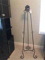 Front view of the black metal easel stand showing three legs, leaf-shaped top ornament, and scroll details.
