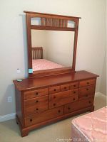 Front view of wooden dresser with attached mirror. Reflects part of a bed frame in background.