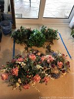 Group of six faux flower arrangements displayed on carpet; two metal-framed baskets with green and white foliage, and four large mixed flower bunches in front with pink roses, blue hydrangeas, and other flowers.