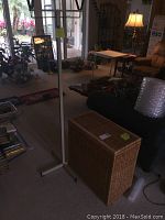 Overall living area view showing the wicker laundry basket positioned near a chair and floor lamp, with the white metal clothing rack standing nearby.