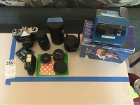 Photo of Polaroid Impulse camera on top of original box, next to Fujica ST705 SLR camera body, assorted Pentax lenses in cases, Vivitar flash, and other accessories on white table.