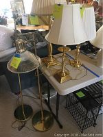 Two adjustable brass floor lamps and three brass table lamps on white table with white shade, cords visible, in light-filled room.