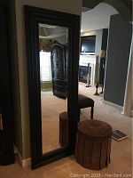 Photo showing the full-length mirror leaning against the wall with the round upholstered stool beside it. Mirror has a dark wooden frame and rectangular shape.