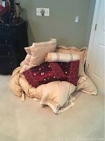 Pile of king size linens and pillows on carpeted floor including cream comforter, various pillows, and a burgundy decorative pillow