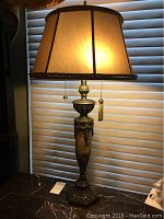 Front view of one decorative table lamp lit showing fabric shade, tassel pull chains, ornate base.