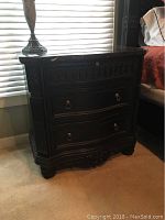 Side view of the wooden side table next to a bed, showing three drawers with metal ring handles and carved details